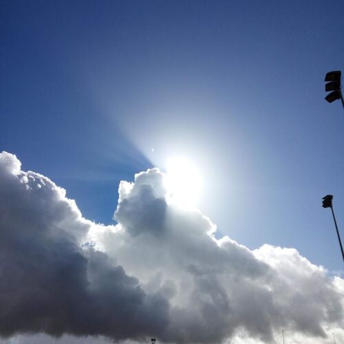Gran nube blanca iluminada por el sol contra un cielo azul, con edificios y farolas al fondo