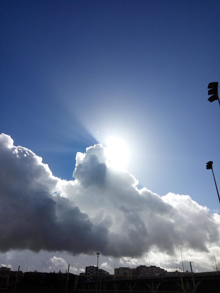 Gran nube blanca iluminada por el sol contra un cielo azul, con edificios y farolas al fondo