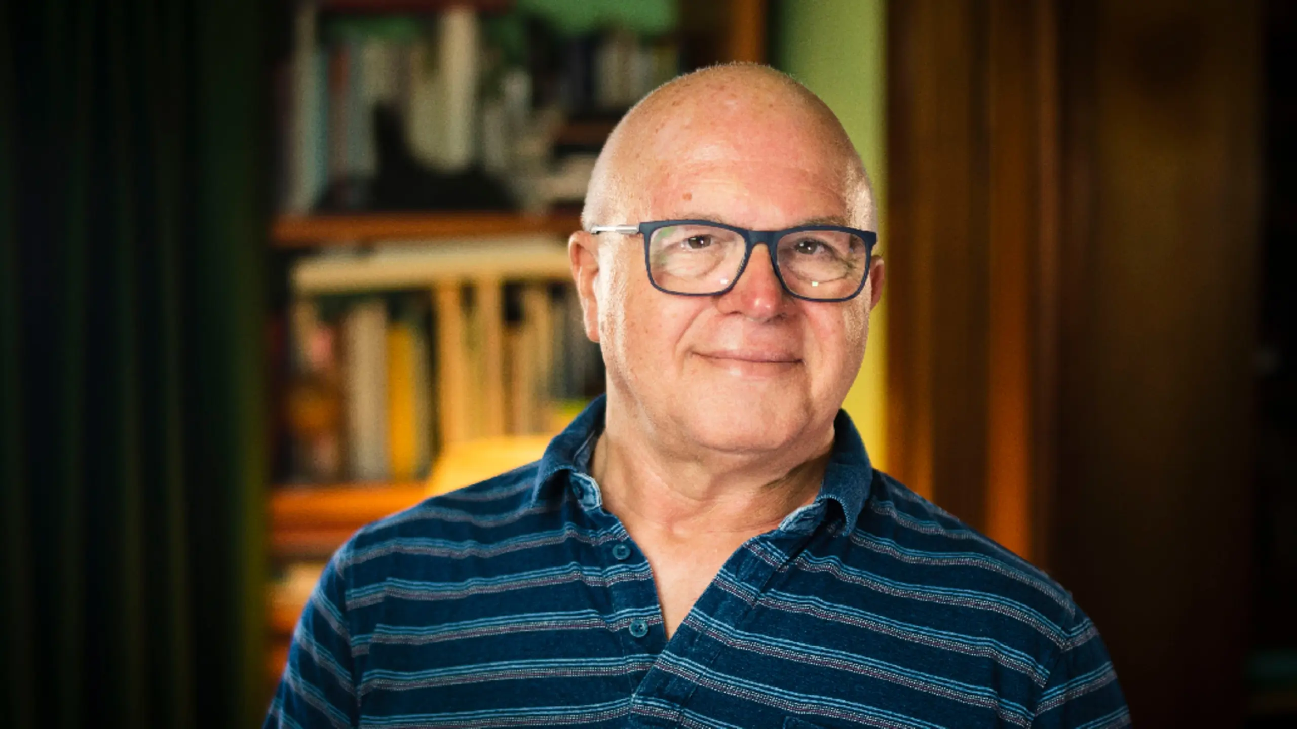 Retrato de Faustino Lobato con gafas y camisa de rayas azules, sonriendo frente a una estantería de libros