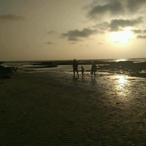 Tres siluetas caminando por la orilla del mar al atardecer, con el sol reflejado en el agua